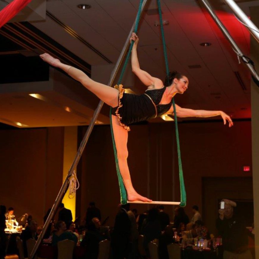 An aerialist from Nimble Arts balances on one leg on a trapeze.
