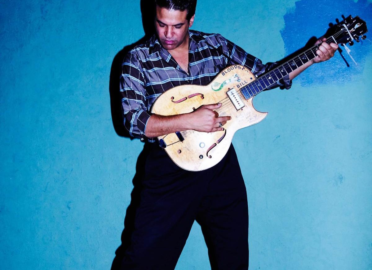 Bloodshot Bill stands in front of a blue wall holding an electric guitar