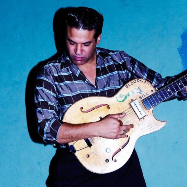 Bloodshot Bill stands in front of a blue wall holding an electric guitar