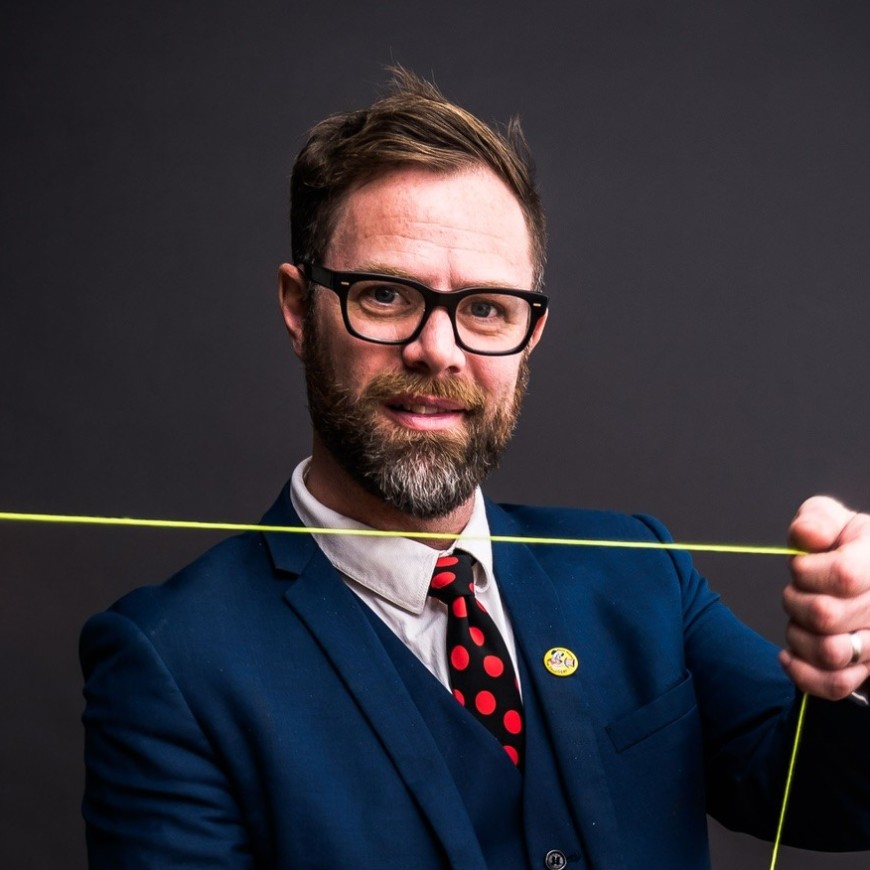 John Higby wears a navy blue suit with a red polka dot tie and spins a yo-yo horizontally.