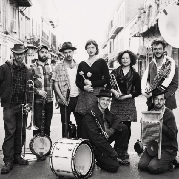 The 8 members of Tuba Skinny stand in the middle of an empty street holding their instruments.