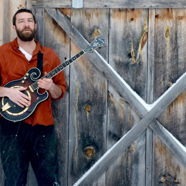 Nate Wallace-Guskov holds a banjo and stands in front of a barn door