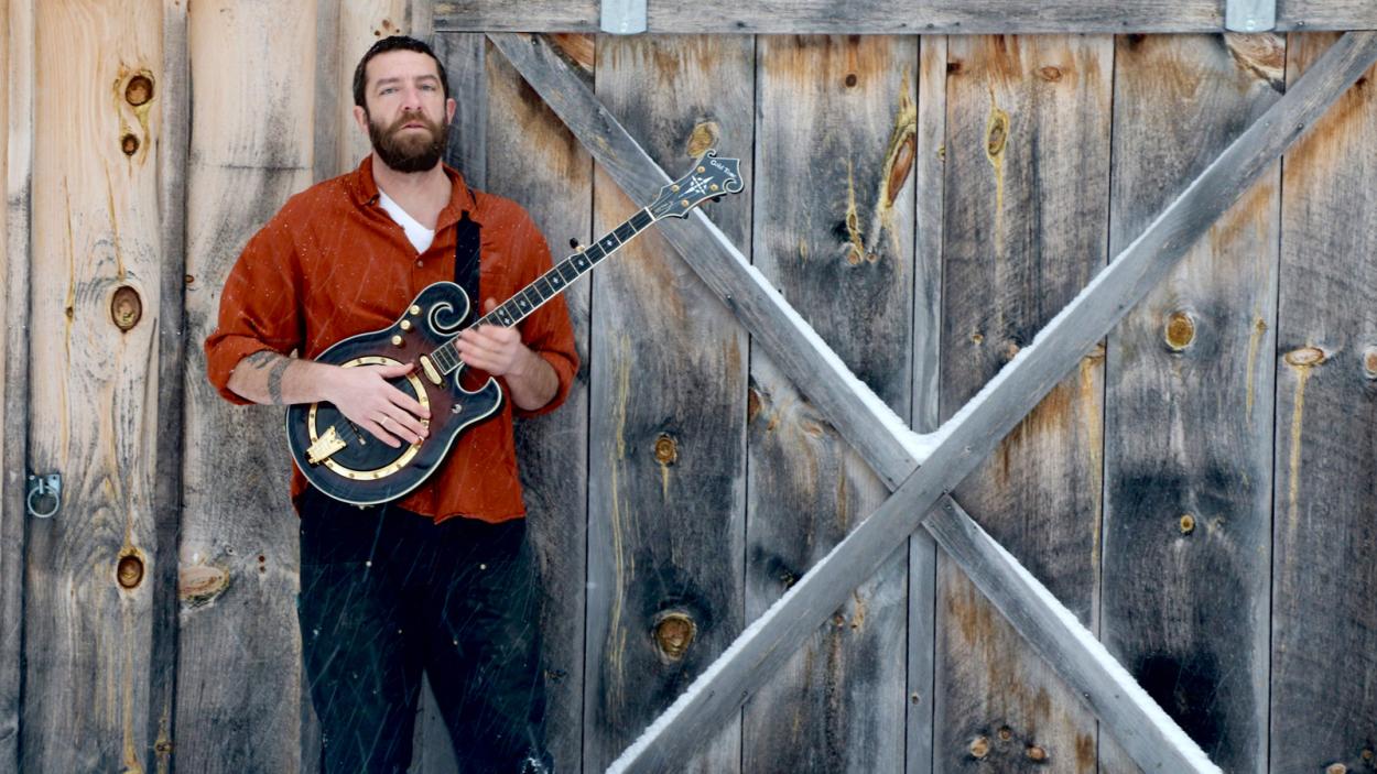 Nate Wallace-Guskov holds a banjo and stands in front of a barn door