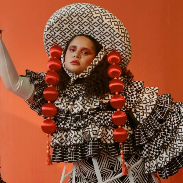 A medium skinned woman with dark hair wears an elaborate black and white ruffled patterned ruffled dress with a matching wide brimmed hat trimmed with two long strands of red balls that hang down beside her face. She stands against an orange backdrop and hold a marionette dressed in a similar style in one hand