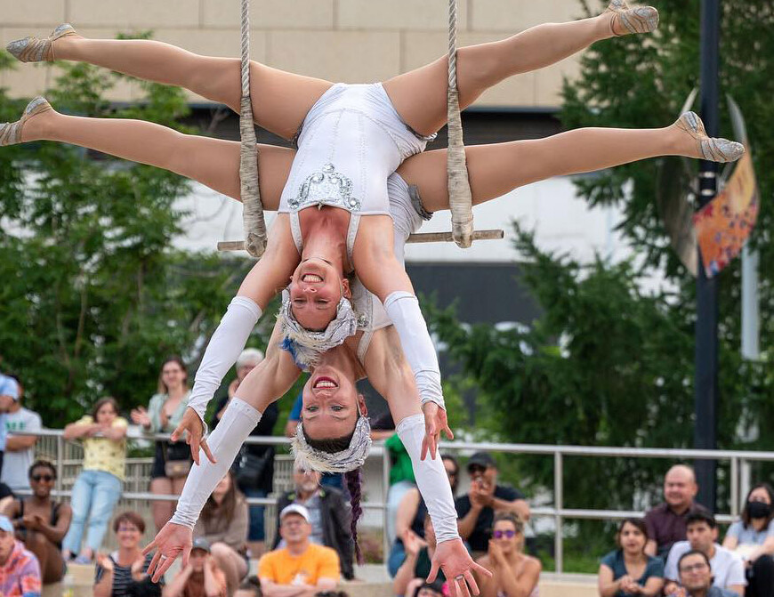 Two light skinned woman hang upside down together from the same trapeze with their legs in a split