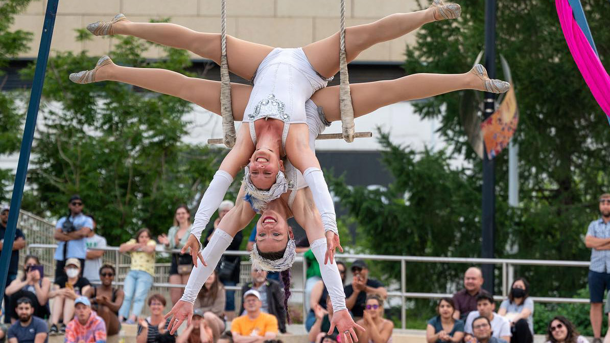 Two light skinned woman hang upside down together from the same trapeze with their legs in a split