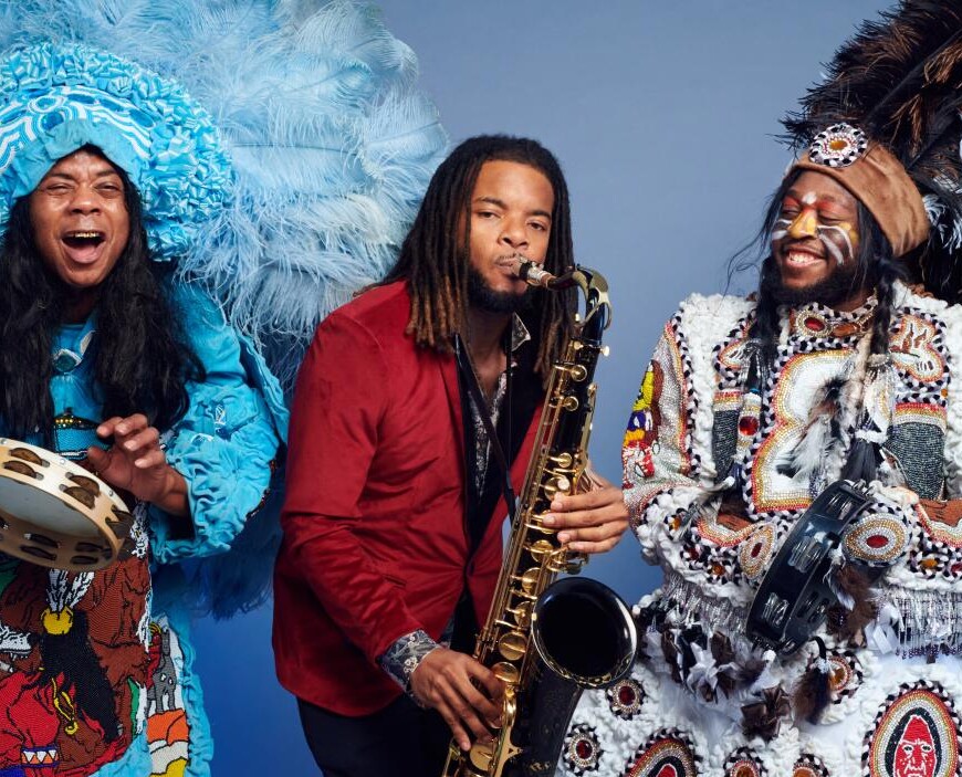 Three dark skinned men play musical instruments against a light blue background. On the right, one man wears a large blue feathered headdress, a blue patterned and ruffled coat, and plays the tambourine and sings. In the middle, one man wears a red blazer and plays the saxophone. On the left one man has orange and yellow stripes painted across his cheeks and wears a large brown, black, and white feathered headdress and a white embroidered and patterned coat while he plays the tambourine