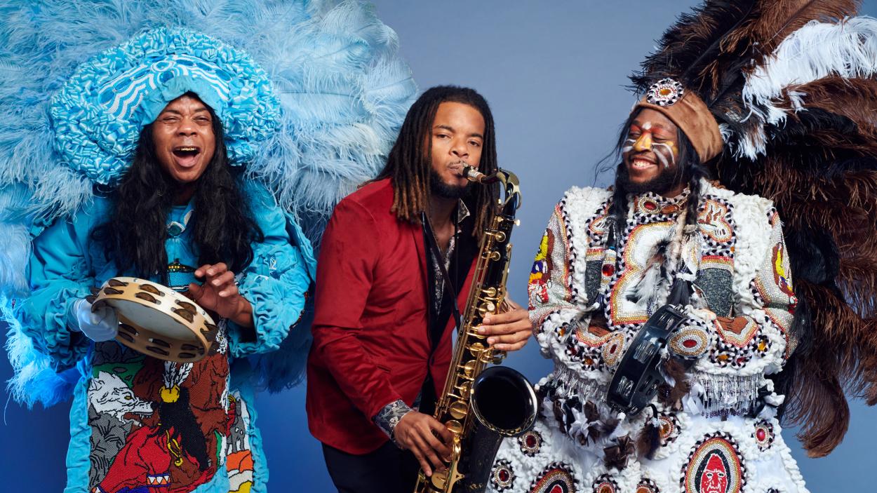 Three dark skinned men play musical instruments against a light blue background. On the right, one man wears a large blue feathered headdress, a blue patterned and ruffled coat, and plays the tambourine and sings. In the middle, one man wears a red blazer and plays the saxophone. On the left one man has orange and yellow stripes painted across his cheeks and wears a large brown, black, and white feathered headdress and a white embroidered and patterned coat while he plays the tambourine