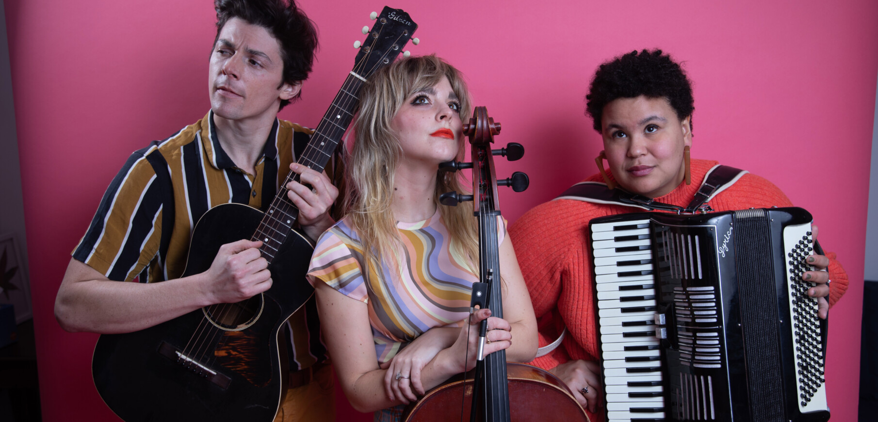 Three people seen from the waist up stand together against a pink background.A light skinned man holds a guitar, a light skinned woman holds a bass, and a dark skinned woman wears an accordian