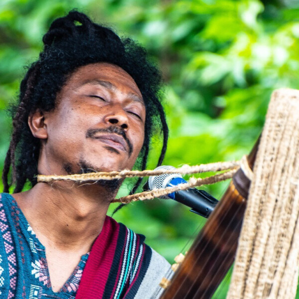 a dark skinned man plays a multi stringed instrument with his eyes closed