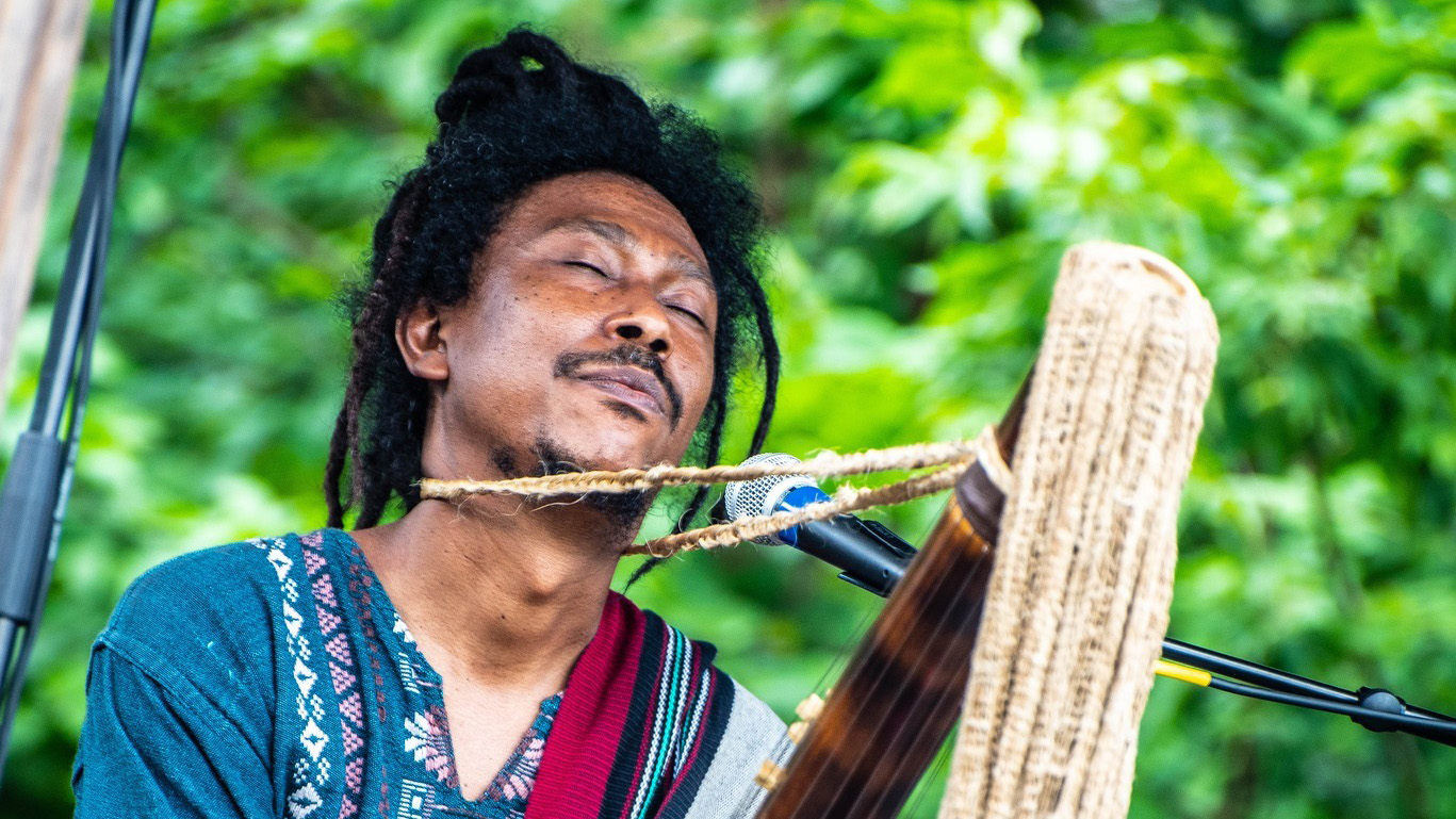 a dark skinned man plays a multi stringed instrument with his eyes closed