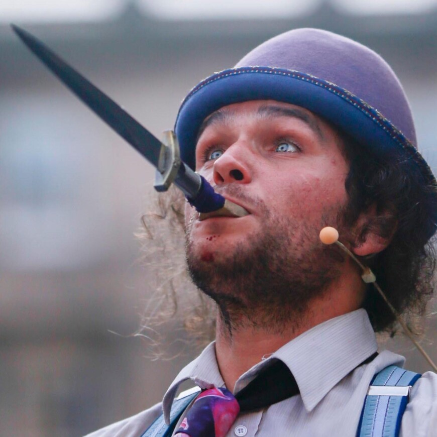 A headshot of a light skinned man wearing a purple bowler hat holding the hilt of a dagger in his mouth