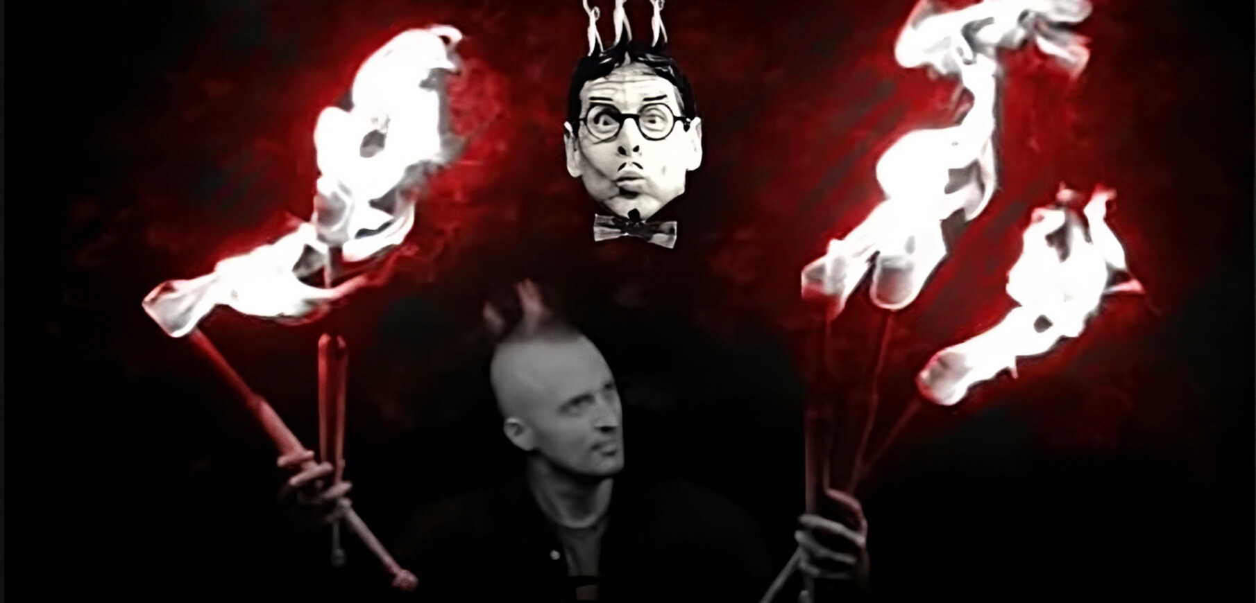 The faces of two light skinned men hover against a dark backdrop. One appears t have glasses and a moustaches drawn on with marker. Hands holding flaming batons frame them.