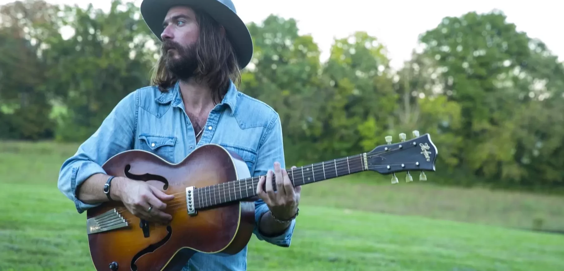 a light skinned man with shoulder length brown hair and a beard wears a flat brimmed hat and holds a guitar as he stands in a green field
