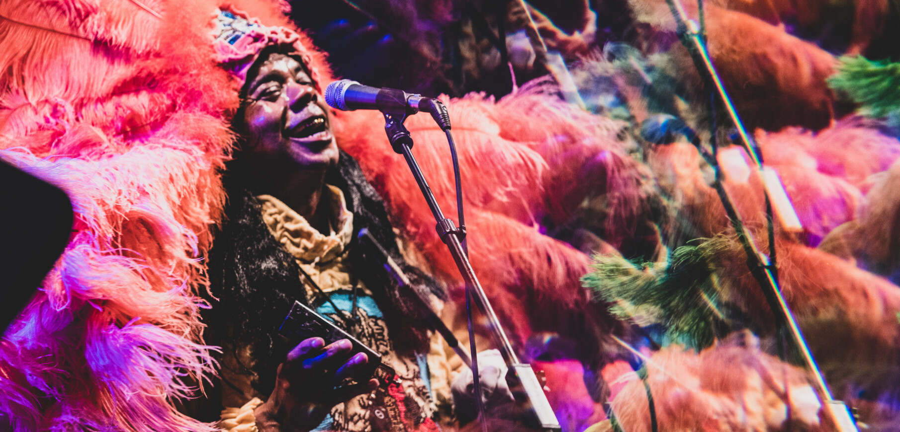a dark skinned man wears a pink, purple, and green feathered headdress and coat and sings into a microphone