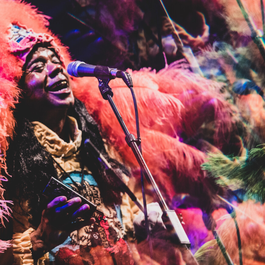 a dark skinned man wears a pink, purple, and green feathered headdress and coat and sings into a microphone