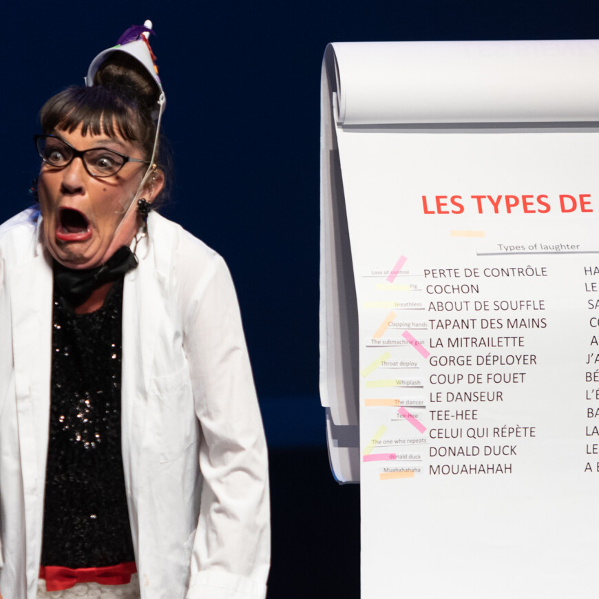 A light skinned woman wears a white lab coat and white plastic funnel as a hat. She makes a surprised face while standing next to a large pad of paper covered with french phrases and post-it notes