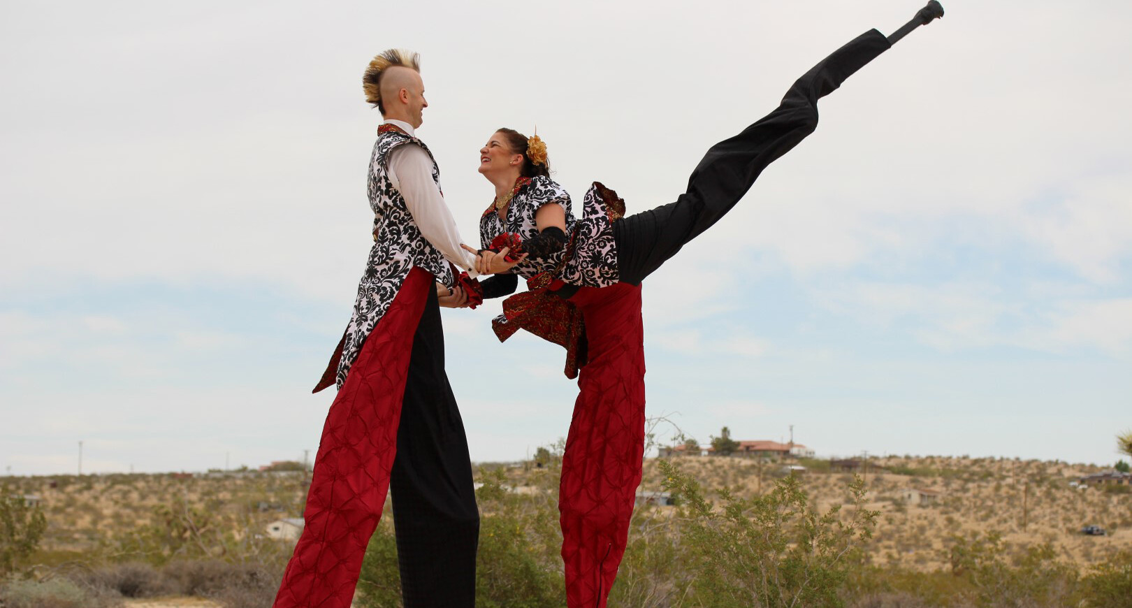A light skinned man and a woman dance together on stilts. Both wear black and white patterned brocade waistcoats with pants that have one black eg and one red