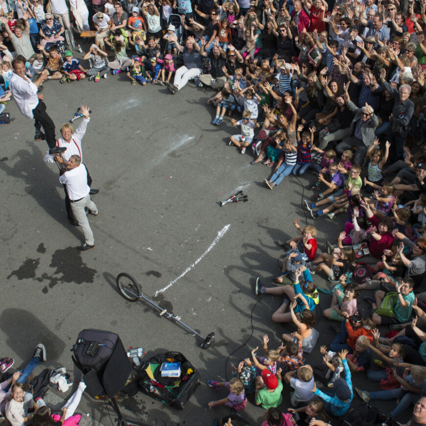 Two light skinned men seen from above on tall unicycles surrounded by a crowd