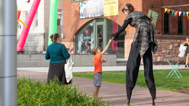 A woman on stilts, seen from behind bends down and hands something to a small boy wearing an orange t shirt a