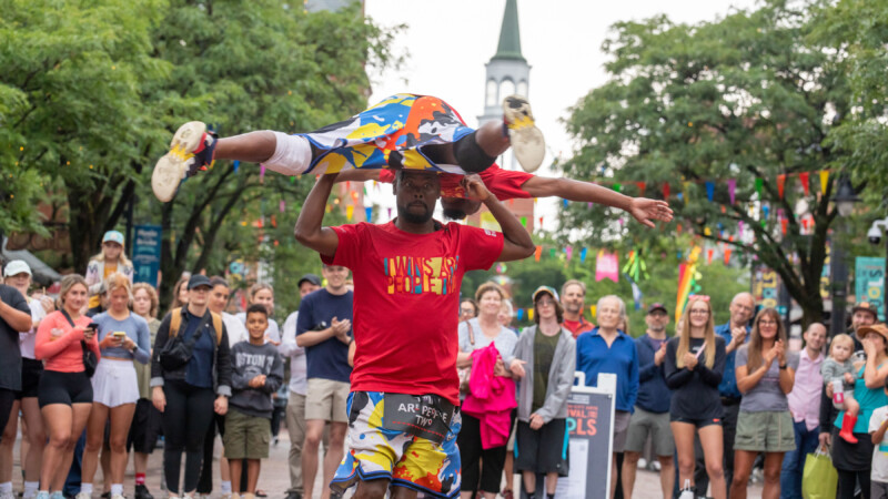 One dark skinned man balances another dark skinned man on his head while a crowd looks on