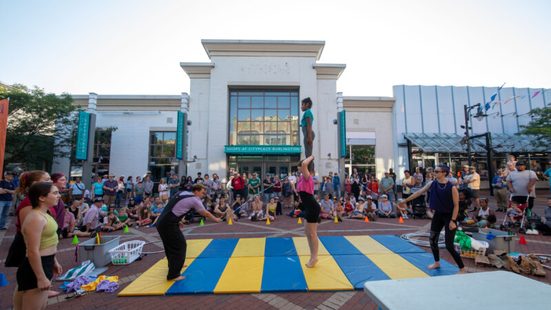 A light skinned man and woman do acrobatic tricks on a blue and yellow striped mat on a brick street as a crowd looks on