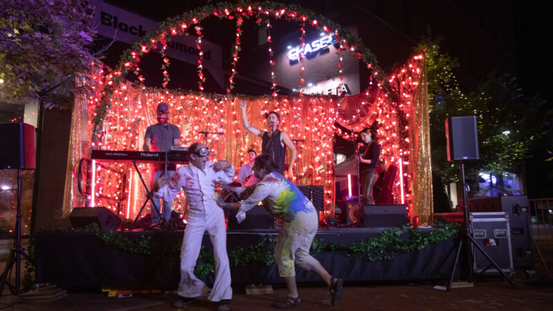 Three light skinned musicians play on a stage backed by range string lights while two people wearing white paint stained jumps suits dance in front