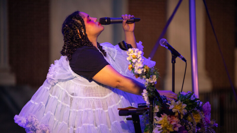 A medium skinned woman wears a tiered blue tulle top and sings into a microphone draped in flowers