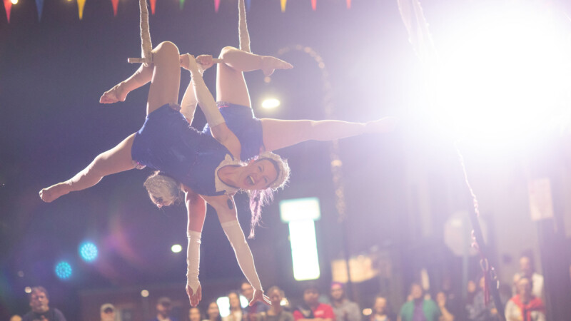 Two light skinned women hang from a trapeze at night, with a bright flash of light beside them