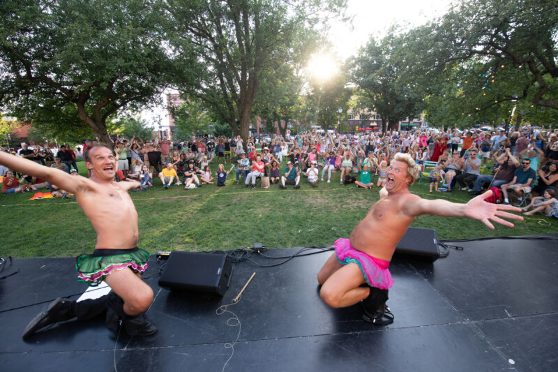 Two light skinned men wear tutus and throw their arms open as they kneel on a black stage with a crowd standing on grass cheering them on