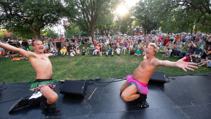 Two light skinned men wear tutus and throw their arms open as they kneel on a black stage with a crowd standing on grass cheering them on