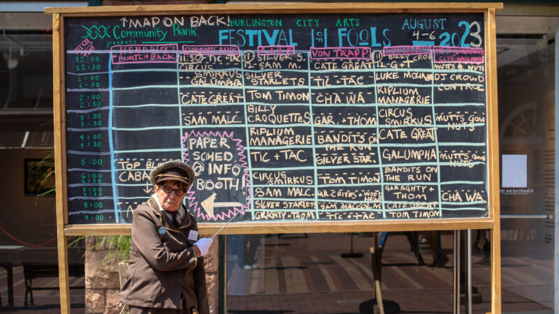 A light skinned woman wearing an old fashioned military uniform stands in front of a chalkboard with a grid schedule written in colorful chalk