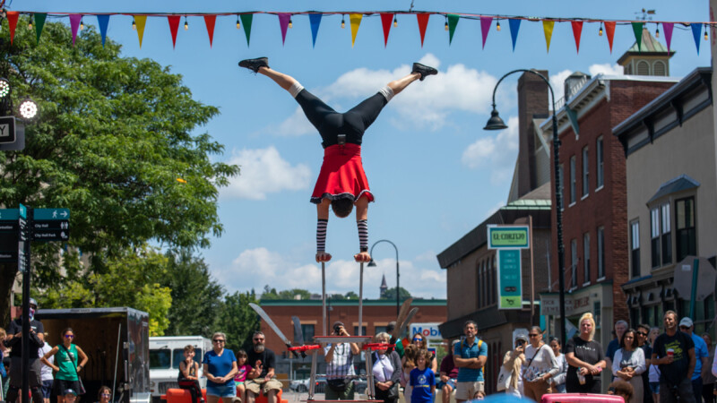 A light skinned woman does a handstand on two poles, seen from behind, as a crowd looks on with blue sky in the background