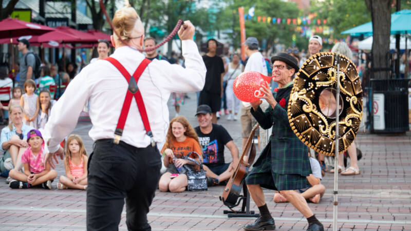 A light skinned man wearing red suspenders seen from behind holds a baton while another light skinned man wearing a green plaid kilt and a black and gold sombrero blows up a red balloon