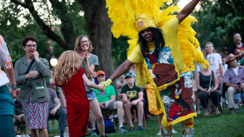 A dark skinned man wearing a yellow feathered headdress and colorful yellow outfit holds hand with a light skinned girl wearing a red dress and dances on the grass in a crowd