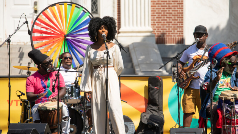 A dark skinned woman and three dark skinned men stand on a stage decorated with rainbow colored whirligigs and sing and play guitar at microphones