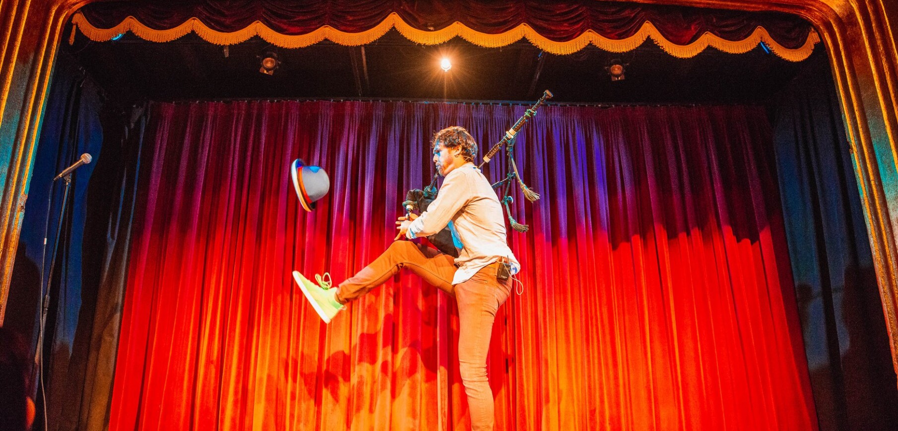 A performer stands on one leg playing the bag pipes and juggling a hat against a red curtain