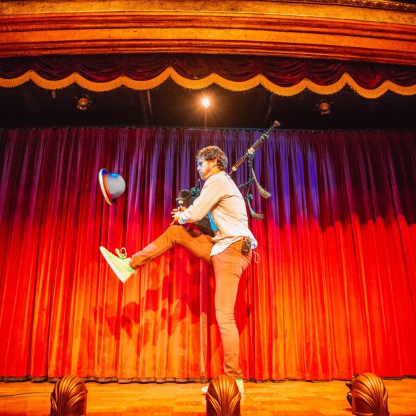A performer stands on one leg playing the bag pipes and juggling a hat against a red curtain