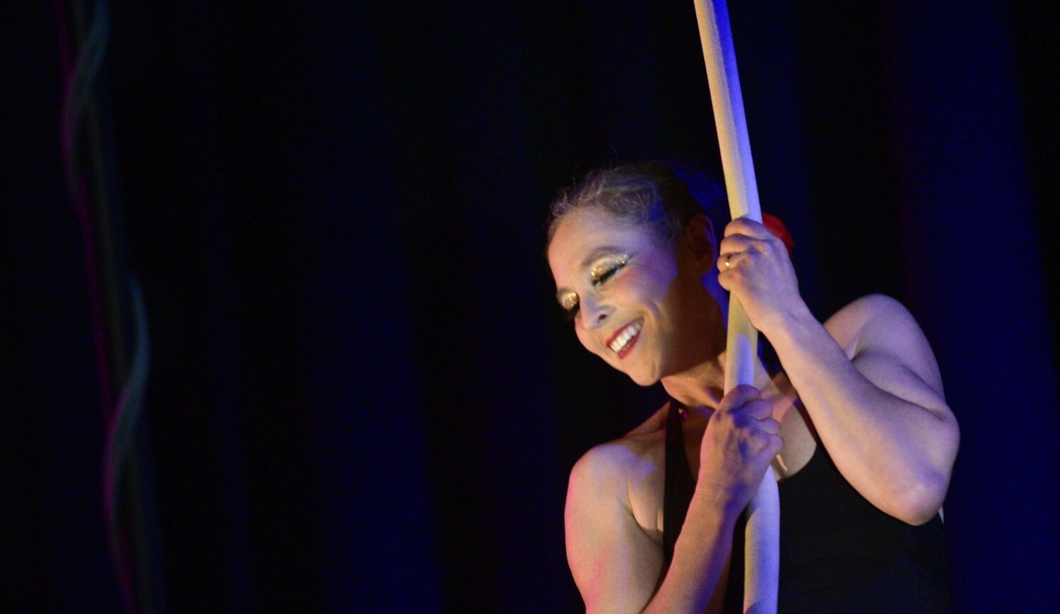 A smiling acrobat holds tight to a rope against a black background.
