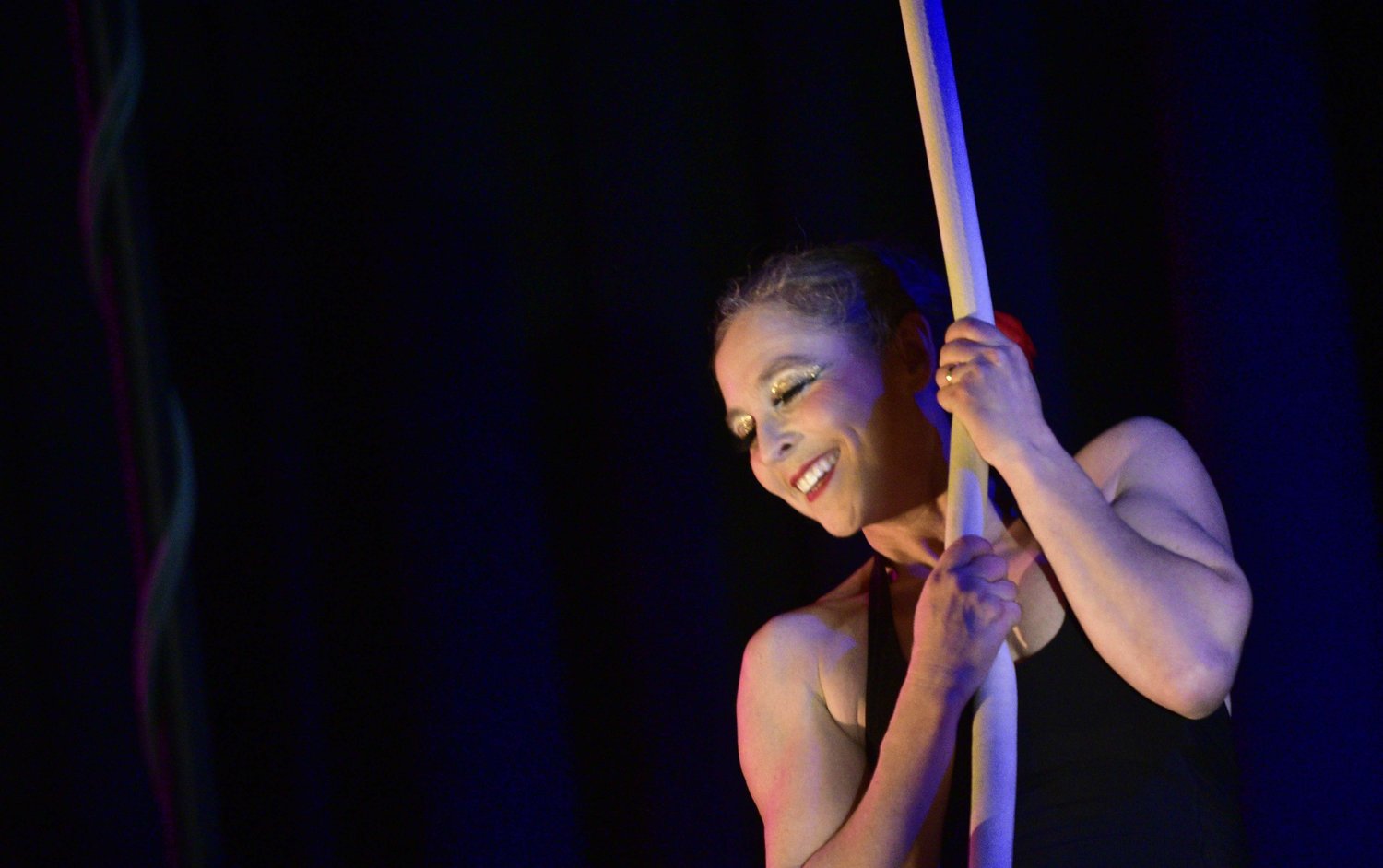 A smiling acrobat holds tight to a rope against a black background.